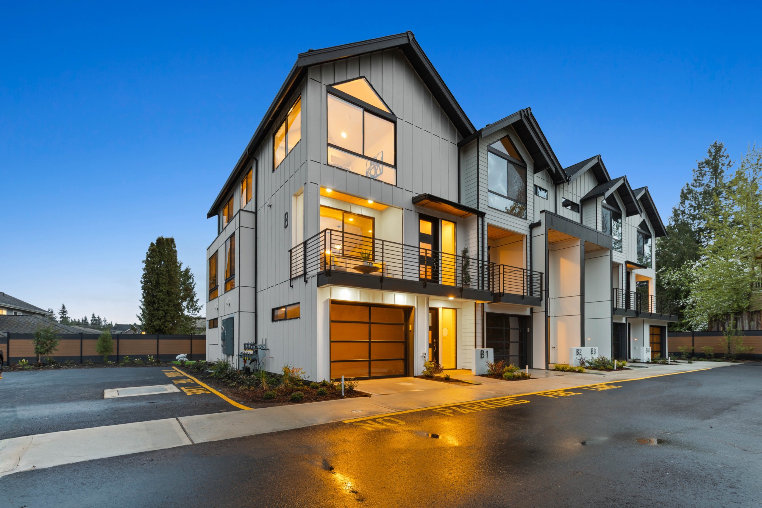 Twilight exterior of a modern luxury townhome with two-car garage, large balconies, warm interior lighting, and contemporary grey and wood facade