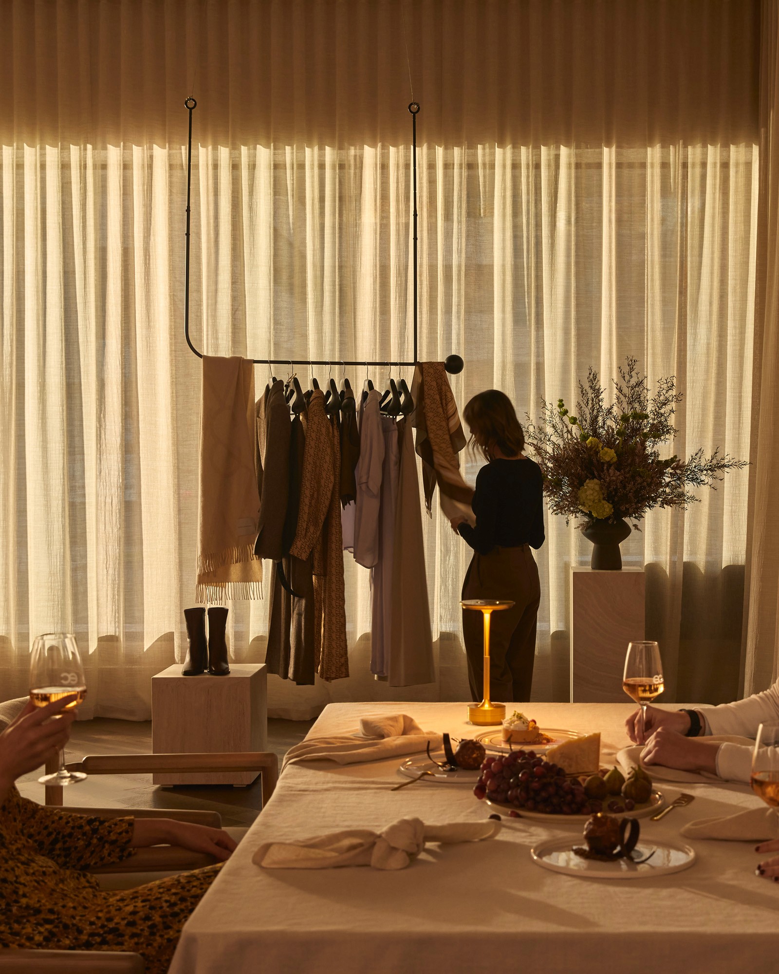 Woman browsing a clothing rack of scarves and garments at a dinner table set with cheese, grapes and figs, wine glasses etched with the eclat logo in the foreground