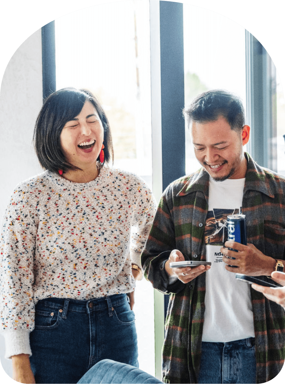 Two coworkers laughing together indoors, one holding a phone and the other holding a drink near large windows.