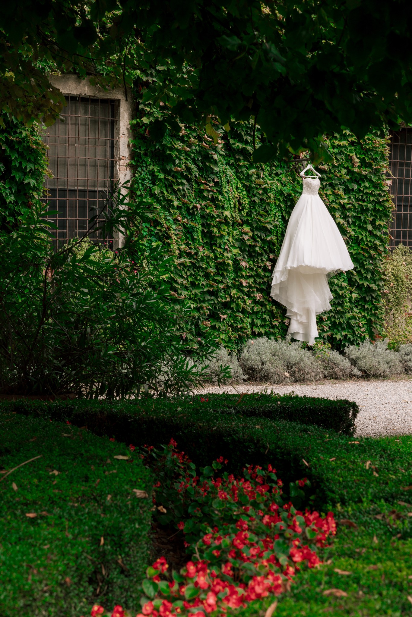 Sposa in abito bianco tra l'edera di Villa Ferrari, preparativi matrimonio in giardino con fiori rossi in primo piano