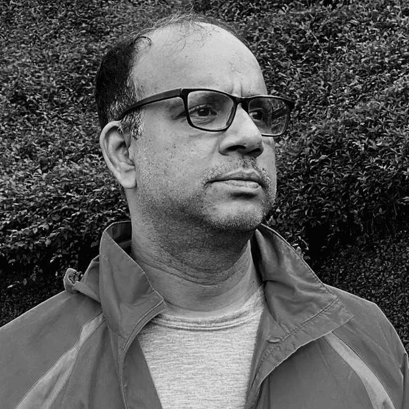 A man reading from a tablet in a collaborative office environment. Desks with documents and a discussion between colleagues are visible in the background.