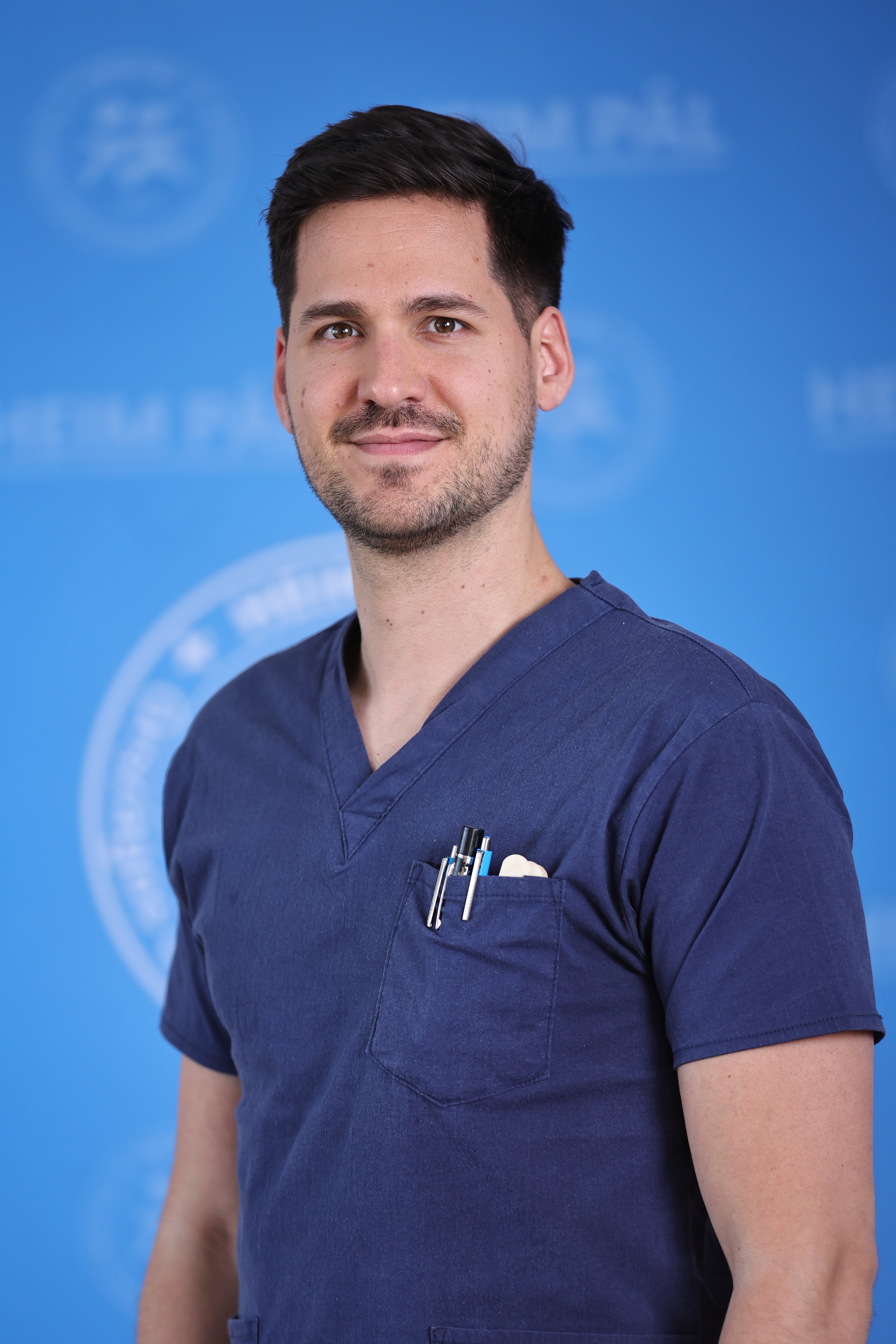 Barna hajú szakállyas férfi bordó orvosi öltözékben sztetoszkóppal kék háttér előtt.