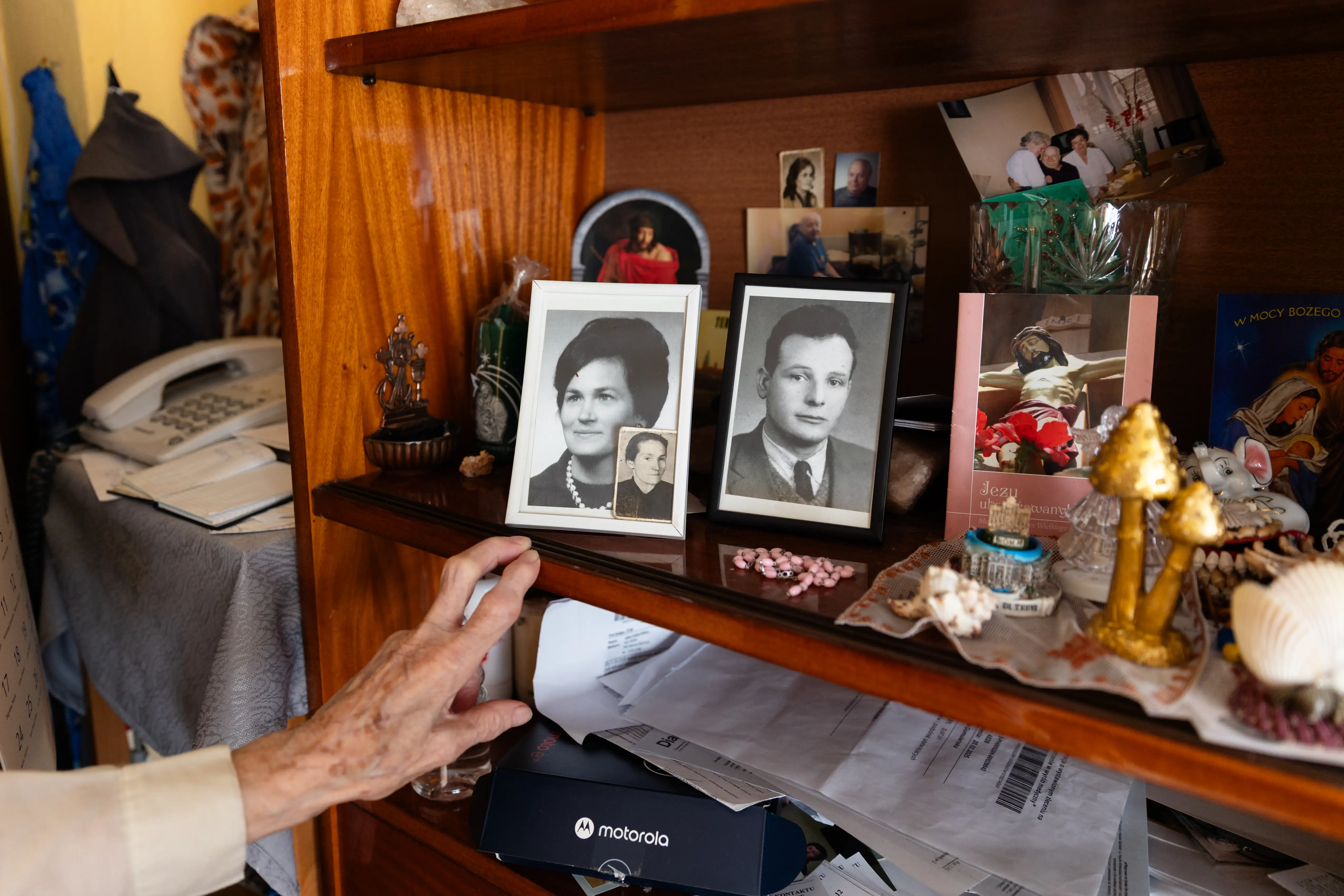 Ręka starszej osoby oparta na półce z pamiątkami. Stoją tam dwa czarno-białe zdjęcia w ramkach, jedno kobiety, a jedno mężczyzny.