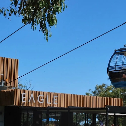 Modern building with "EAGLE" sign and a cable car passing overhead against a clear blue sky. Trees partially visible.