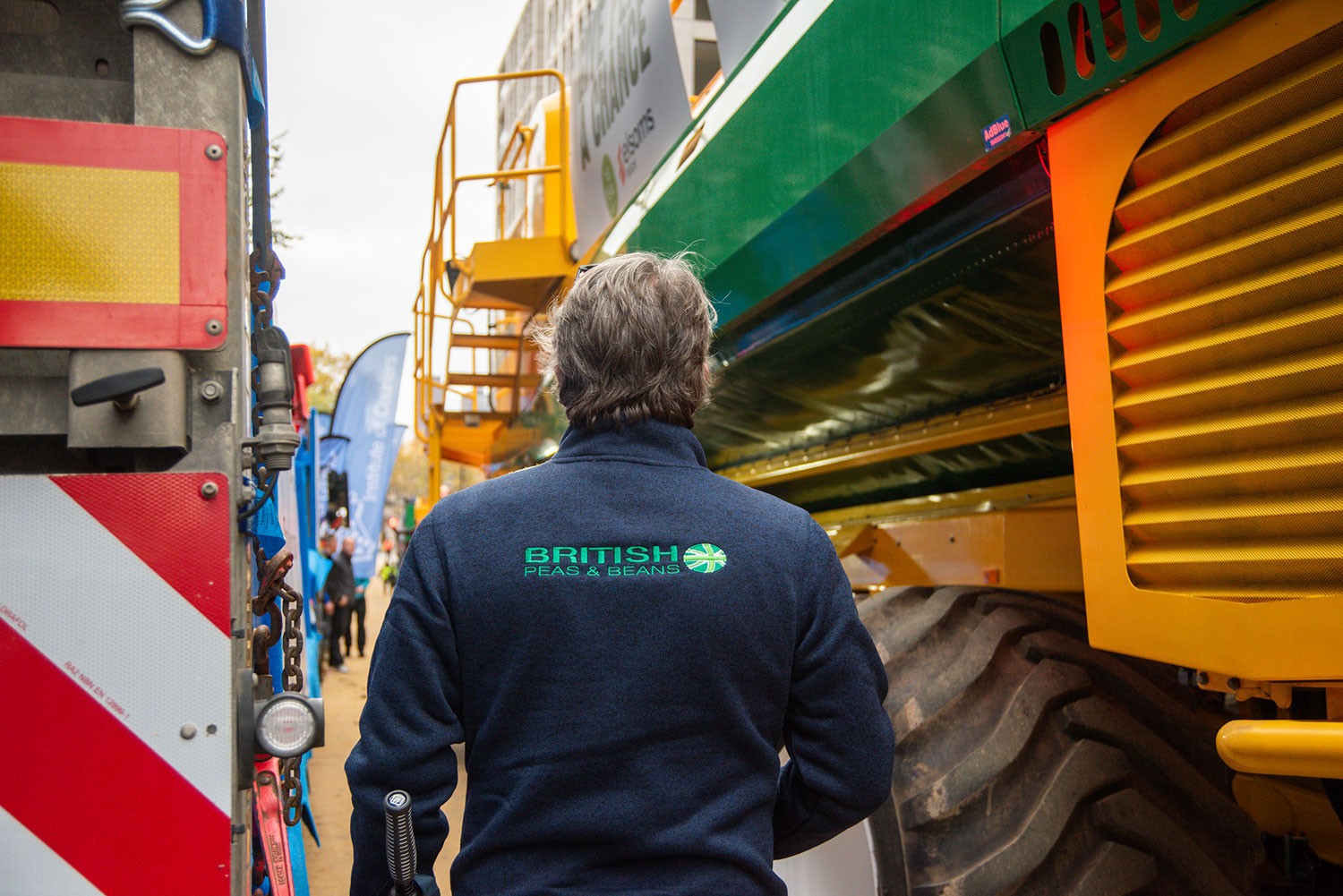 A person wearing a British Peas & Beans branded jacket walks between large pieces of harvesting machinery at an agricultural show.