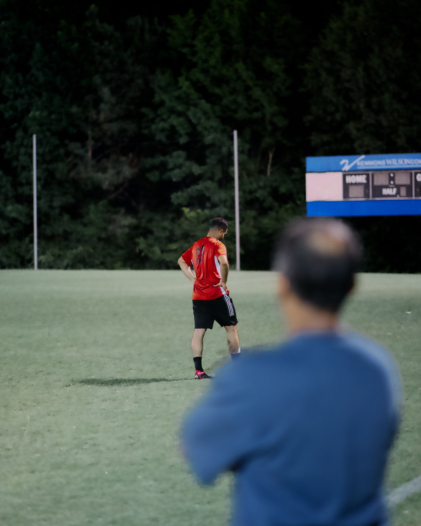 Spectator in the foreground watches an AlFursan FC player near the scoreboard