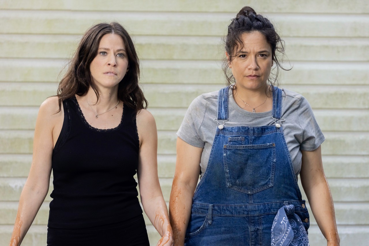 Still from The Floaters: Two women, one in a black tank top and the other in denim overalls, walk side by side with serious expressions.