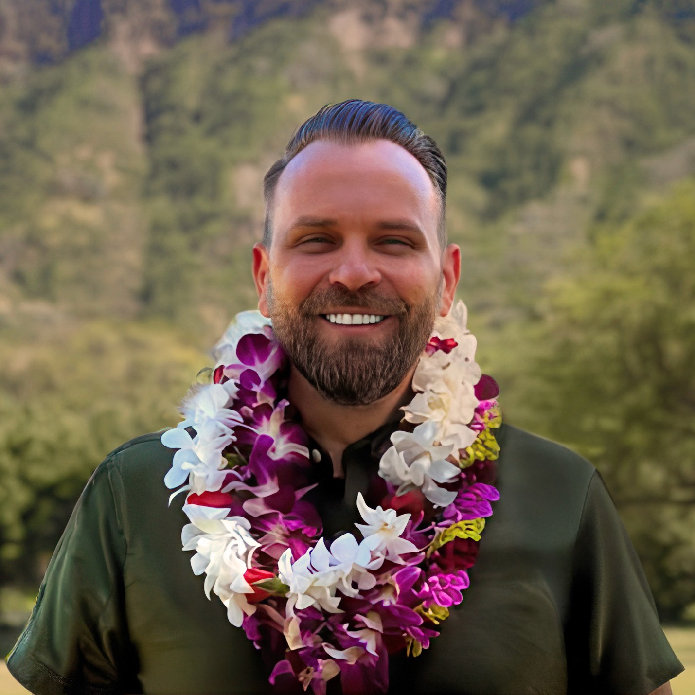 Kevin Welch stands barefoot at the shoreline in Waikiki holding a surfboard and looking toward a vivid ocean sunset after finishing a day of surfing with friends, with gentle waves and distant swimmers in the water and beachfront buildings glowing in the evening light.