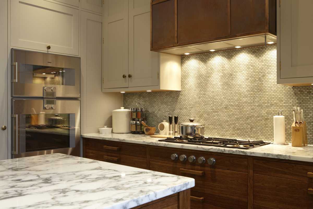 Modern luxury kitchen designed by Doll & Co. City of London Architects featuring marble countertops, mosaic backsplash, warm under-cabinet lighting, and integrated wall ovens.