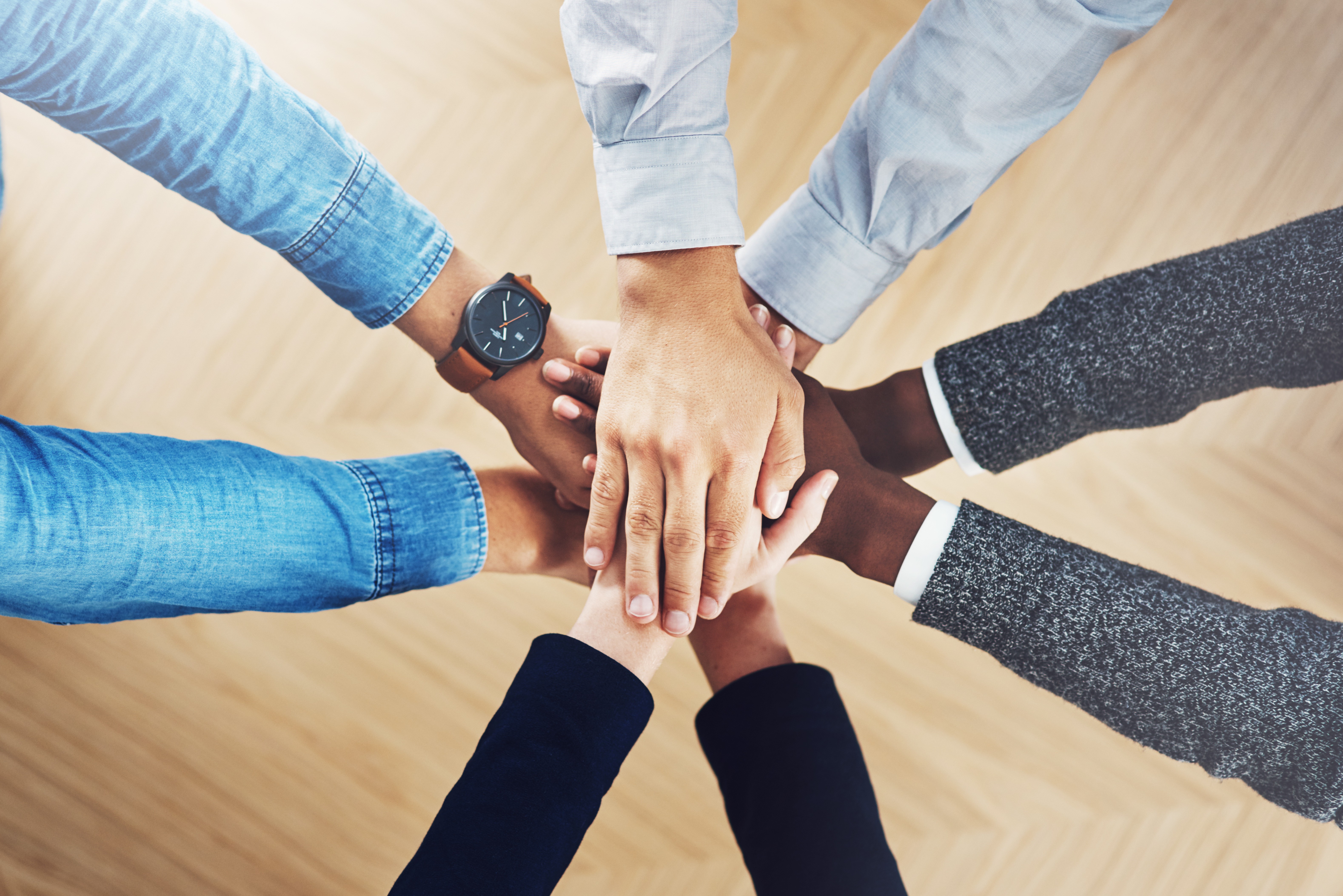 Overhead view of five people with diverse skin tones standing in a circle and stacking their hands together in the center, symbolizing teamwork and collaboration. One person wears a watch and denim shirt, while others wear business-casual clothing, against a light wood floor background.