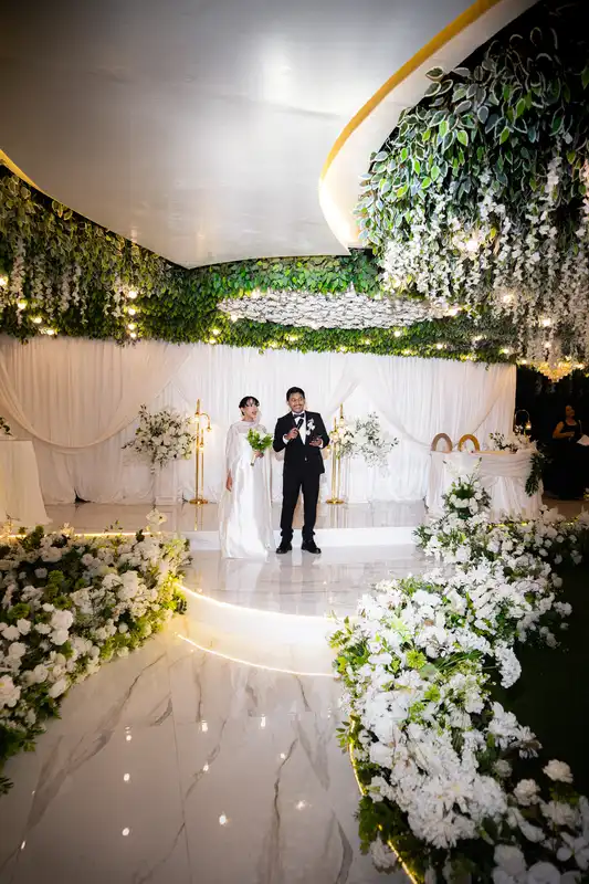 Pasangan pengantin di panggung resepsi putih dengan rangkaian bunga dan drapery elegan