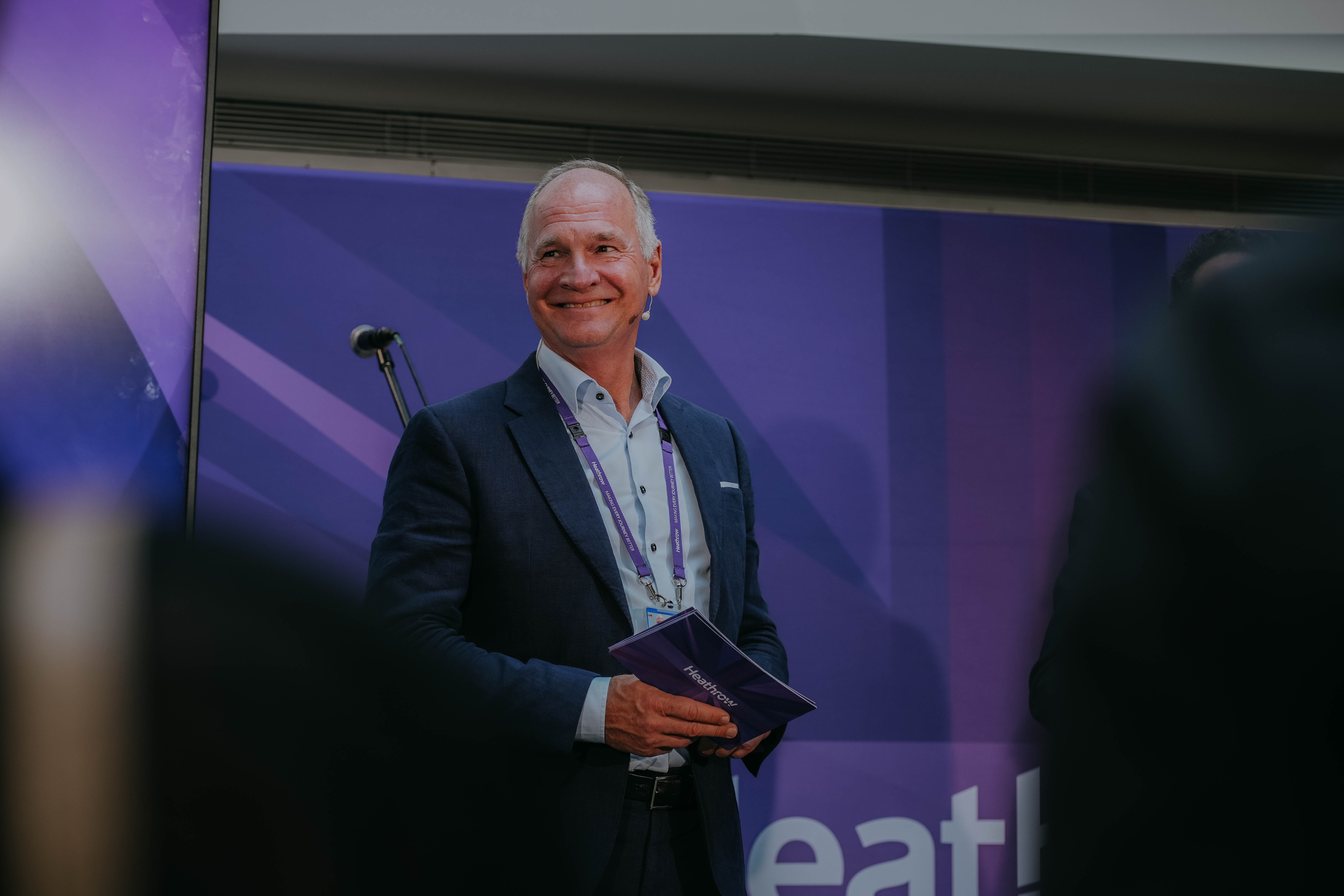 Heathrow CEO Thomas Woldbye smiles warmly while standing on a stage in a dark suit. He is holding a purple pamphlet that says "Heathrow" and is positioned in front of a purple banner.