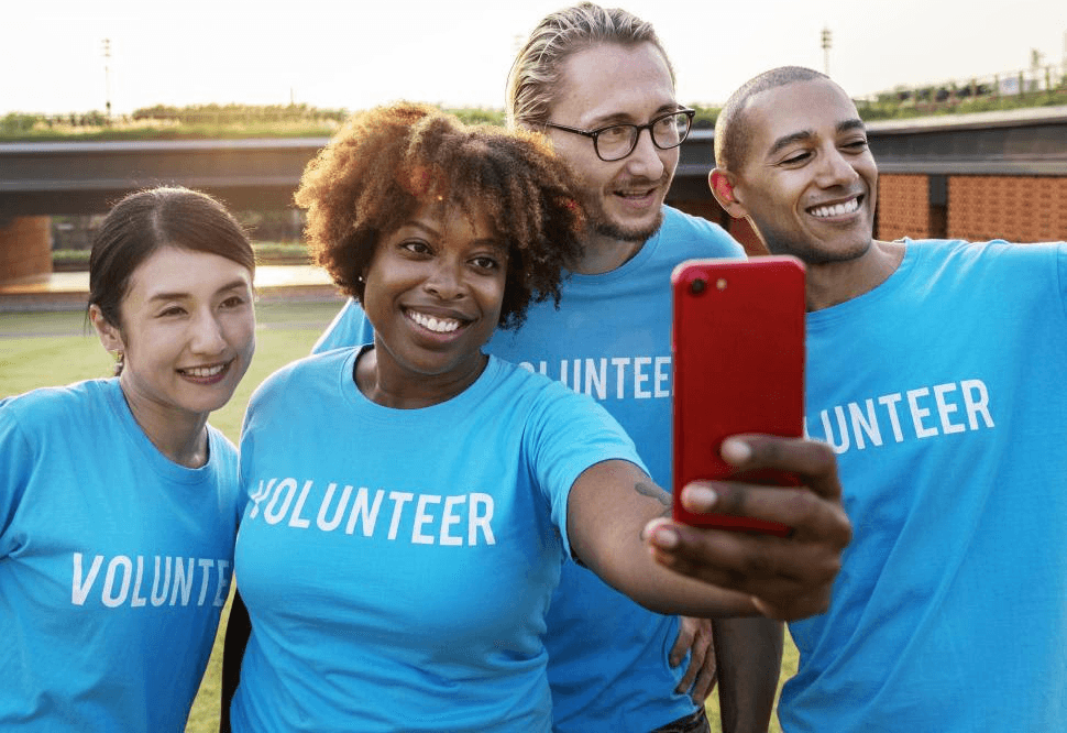 Four friends smiling wearing light blue "volunteer" t-shirts outdoors while one takes a selfie of the group on a red iPhone.