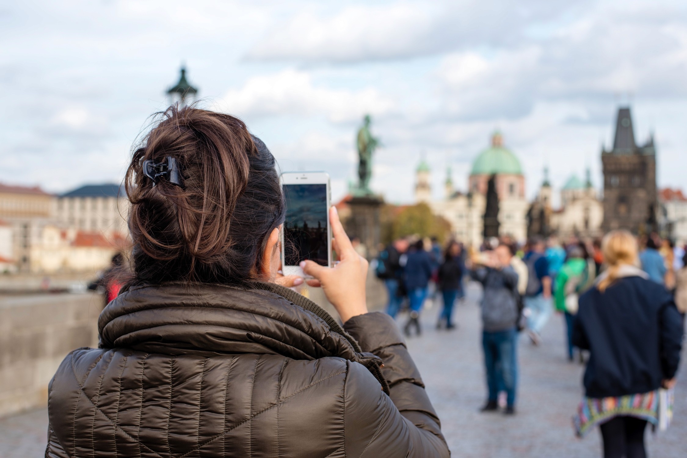 A person takes a picture of distant historic buildings under an overcast sky from a wide, open stone plaza.