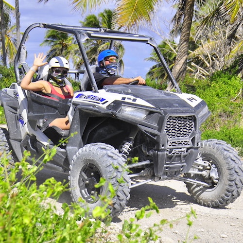 Dos personas con cascos y máscaras montan un buggy en un camino arenoso rodeado de palmeras. Una persona saluda con la mano.