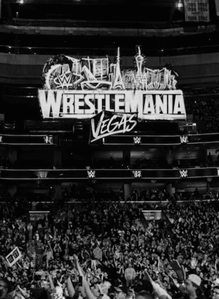 A large crowd fills an arena beneath a bright WrestleMania Las Vegas sign, decorated with iconic Las Vegas landmarks. The scene is lively, with people cheering and holding signs.