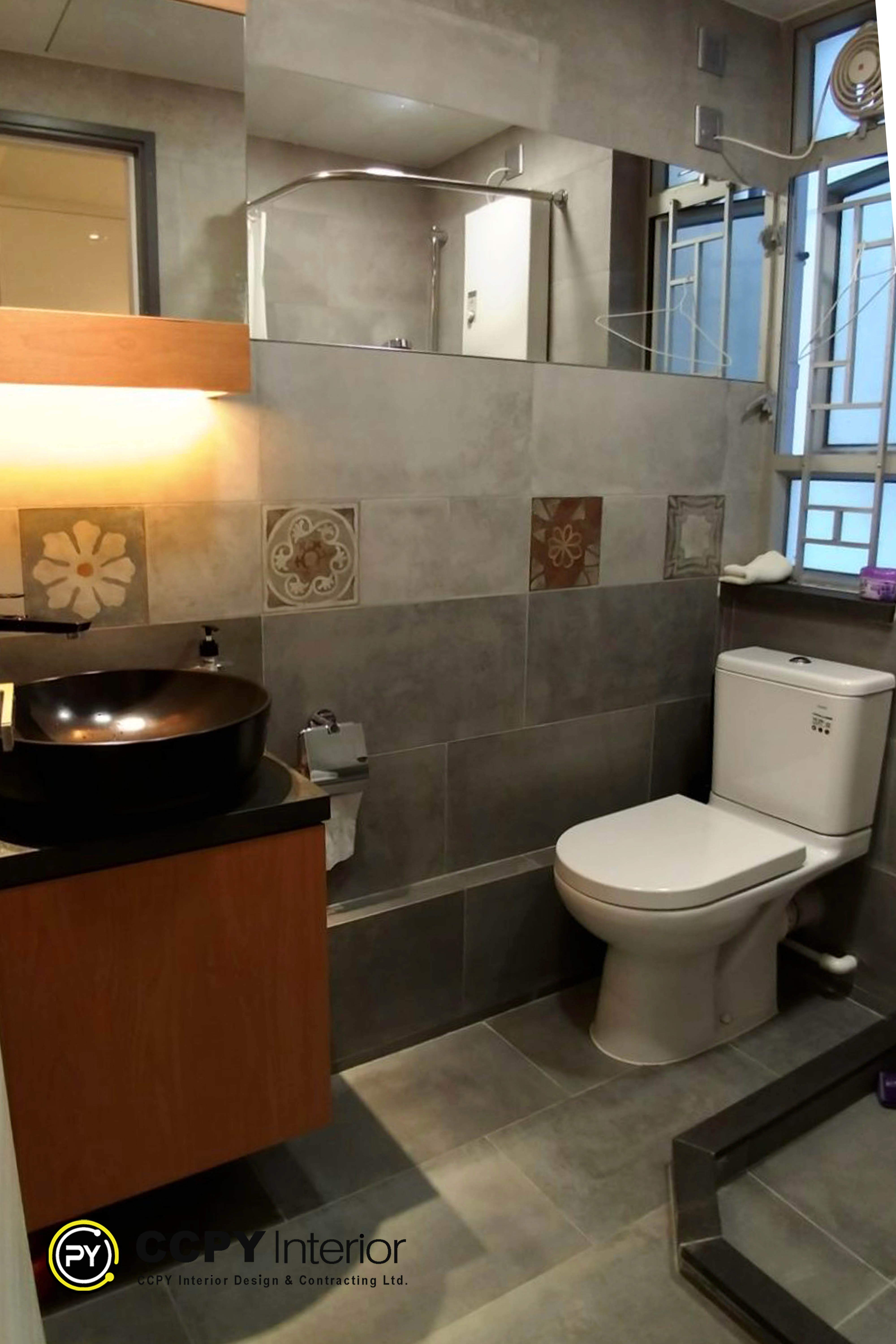 The selected region showcases a modern bathroom renovation featuring neutral grey wall tiling, an artistic bronze vessel sink, and an integrated toilet.Key Design FeaturesColor Palette: Matte grey stone-look wall tiles paired with dark wood cabinetry.Vessel Sink: A distinctive, round metallic bronze basin sitting on a wooden countertop.Accent Tiles: Intricate, square patterned decorative tiles embedded as a feature strip.Lighting: Warm, ambient lighting casting a soft glow above the vanity area.Property
