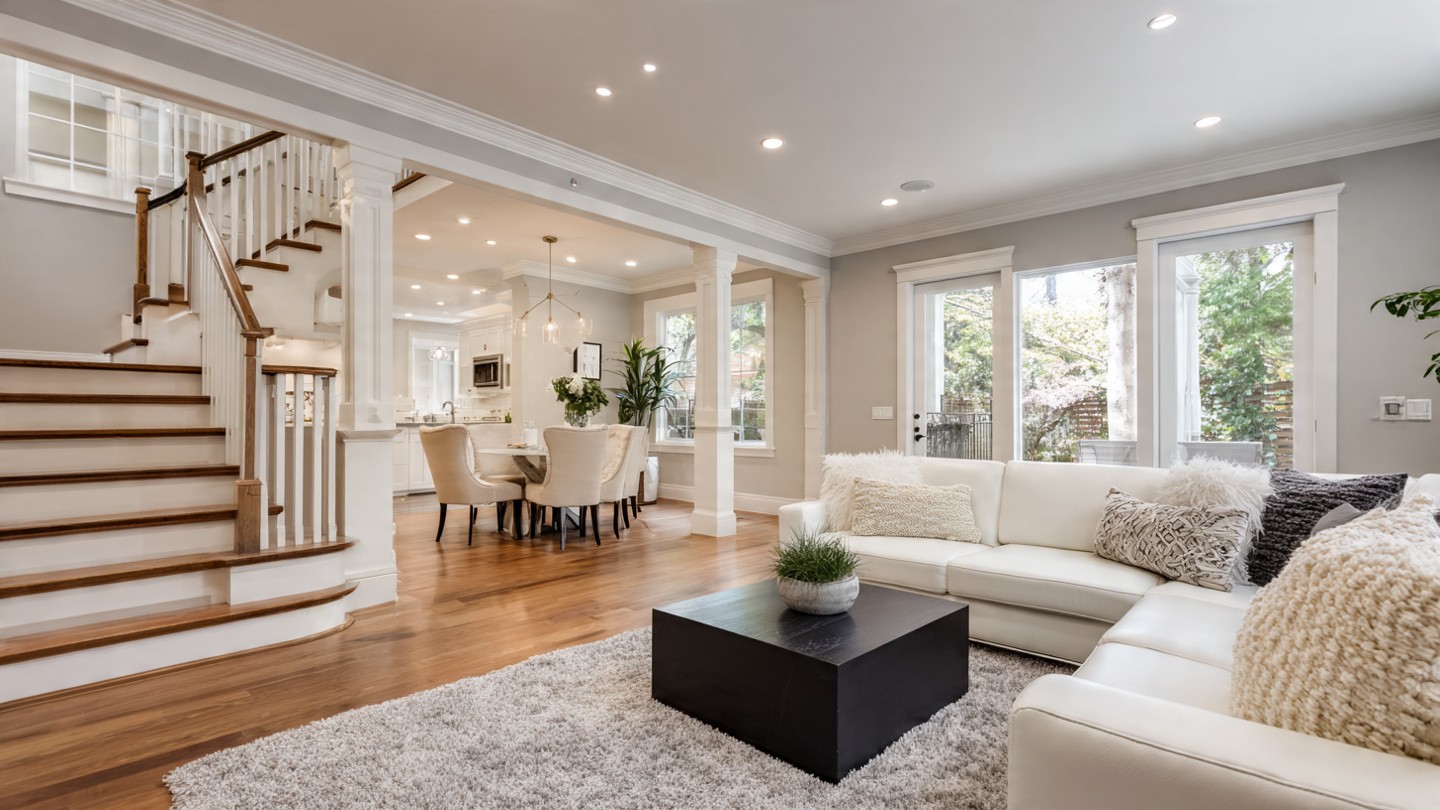Spacious open-concept living room with white sectional sofa, fluffy pillows, and a gray rug. Adjacent dining area, modern kitchen, and wooden staircase.