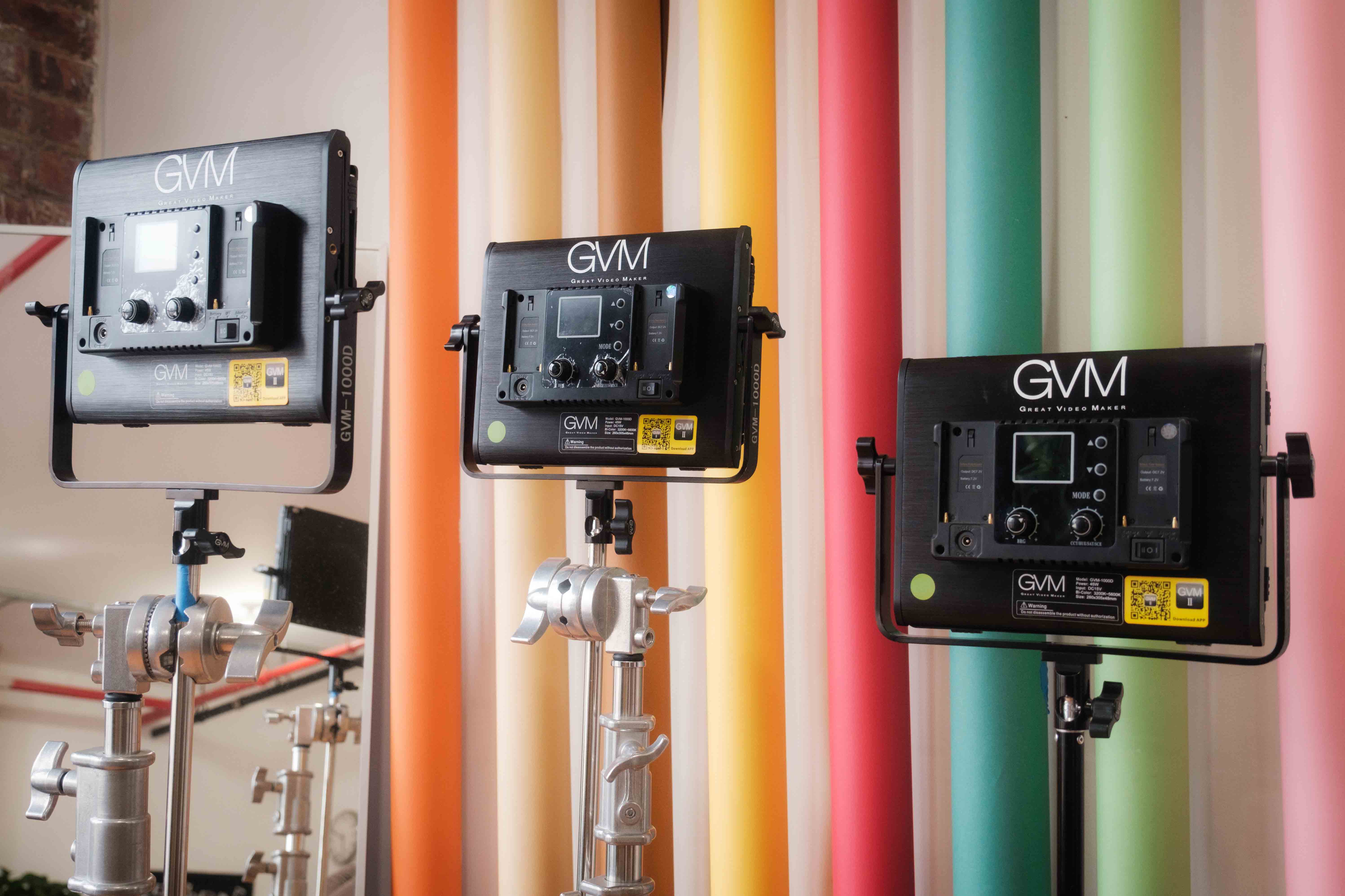 Three GVM video lights on stands in a photo studio, set against a backdrop of colorful paper rolls. The lights are positioned for use, showcasing their digital controls and professional setup.