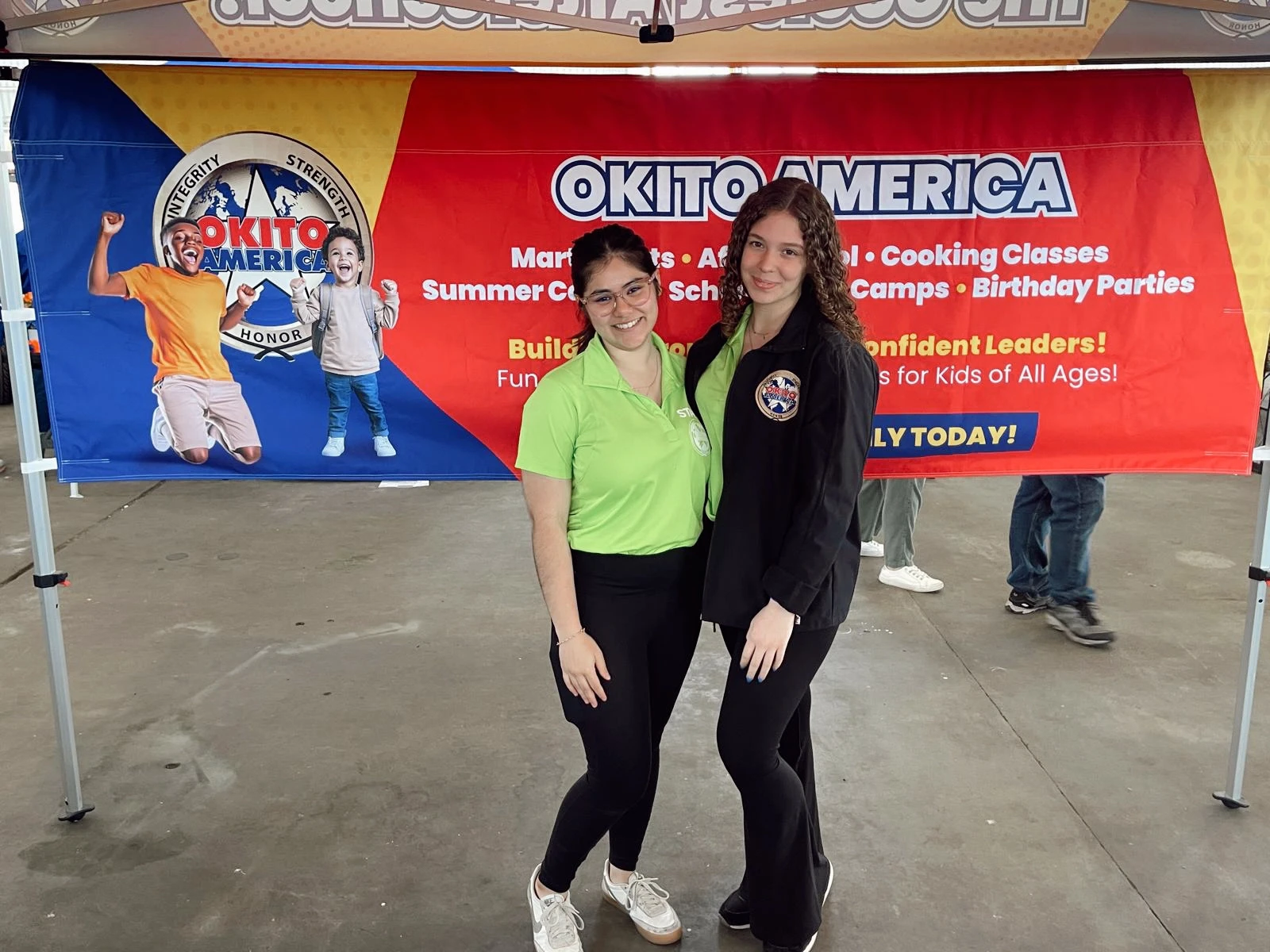 Two female team member standing in front of a Okito banner at an event