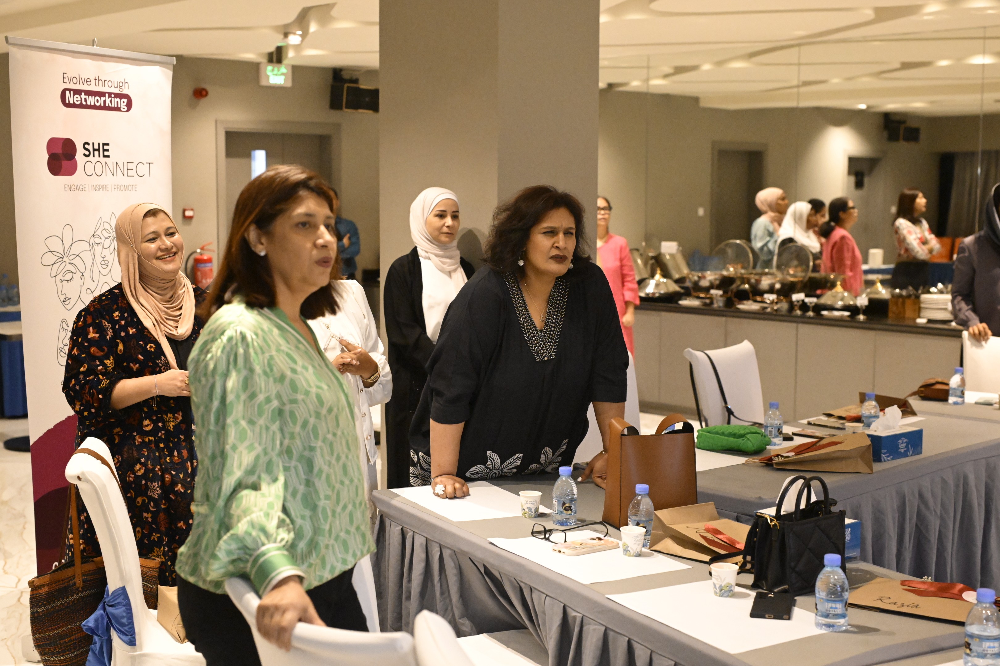 Smiling young woman with long hair standing against a dark green background, holding a finger to her chin.