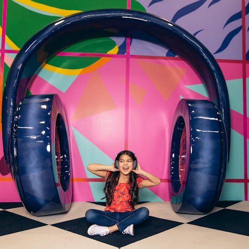 A girl sits cross-legged, smiling with her hands on her cheeks, under large, blue headphone sculptures against a colorful, patterned background.