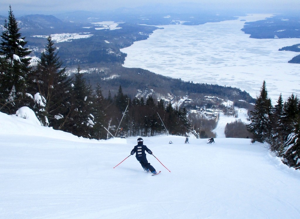 Skieur en descente sur une large piste avec vue panoramique sur le lac Memphrémagog enneigé et les montagnes.