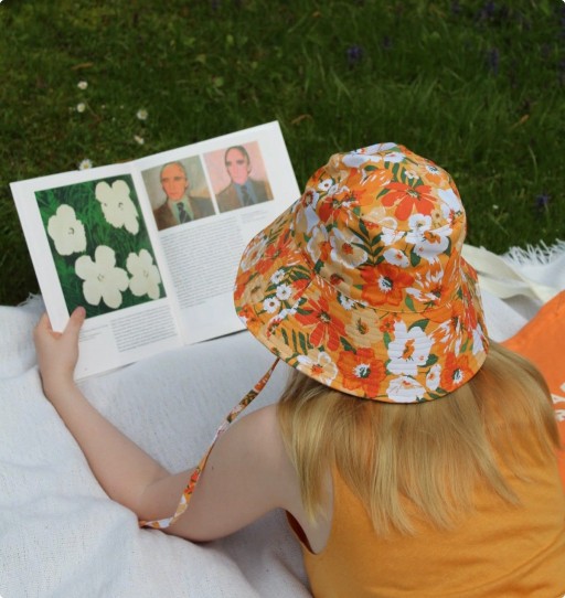 Person on a picnic blanket reading an art book, wearing an orange floral bucket hat on the grass.
