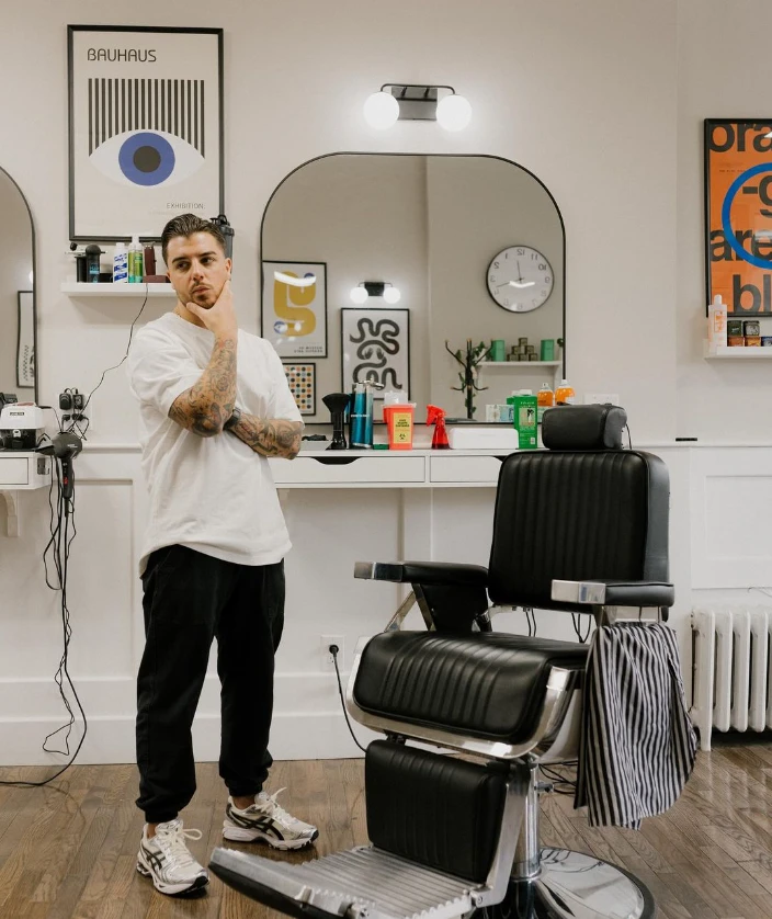 Dom wearing a white t shirt and crossing his arms standing next to a barber chair