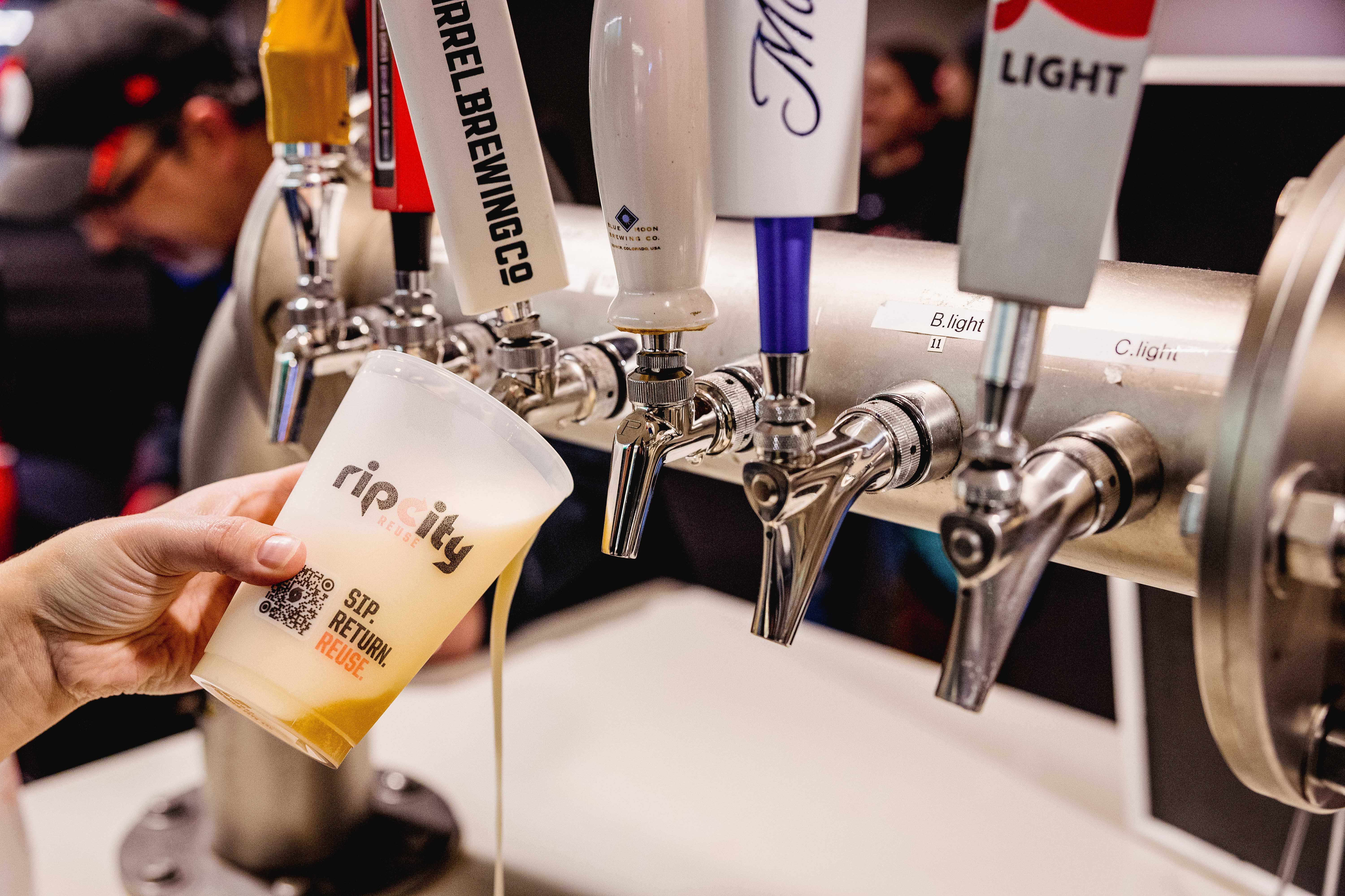 Hand holding a Rip City Reuse cup filled with beer beneath a row of taproom beer taps, with the 'Sip. Return. Reuse.' QR code visible on the cup