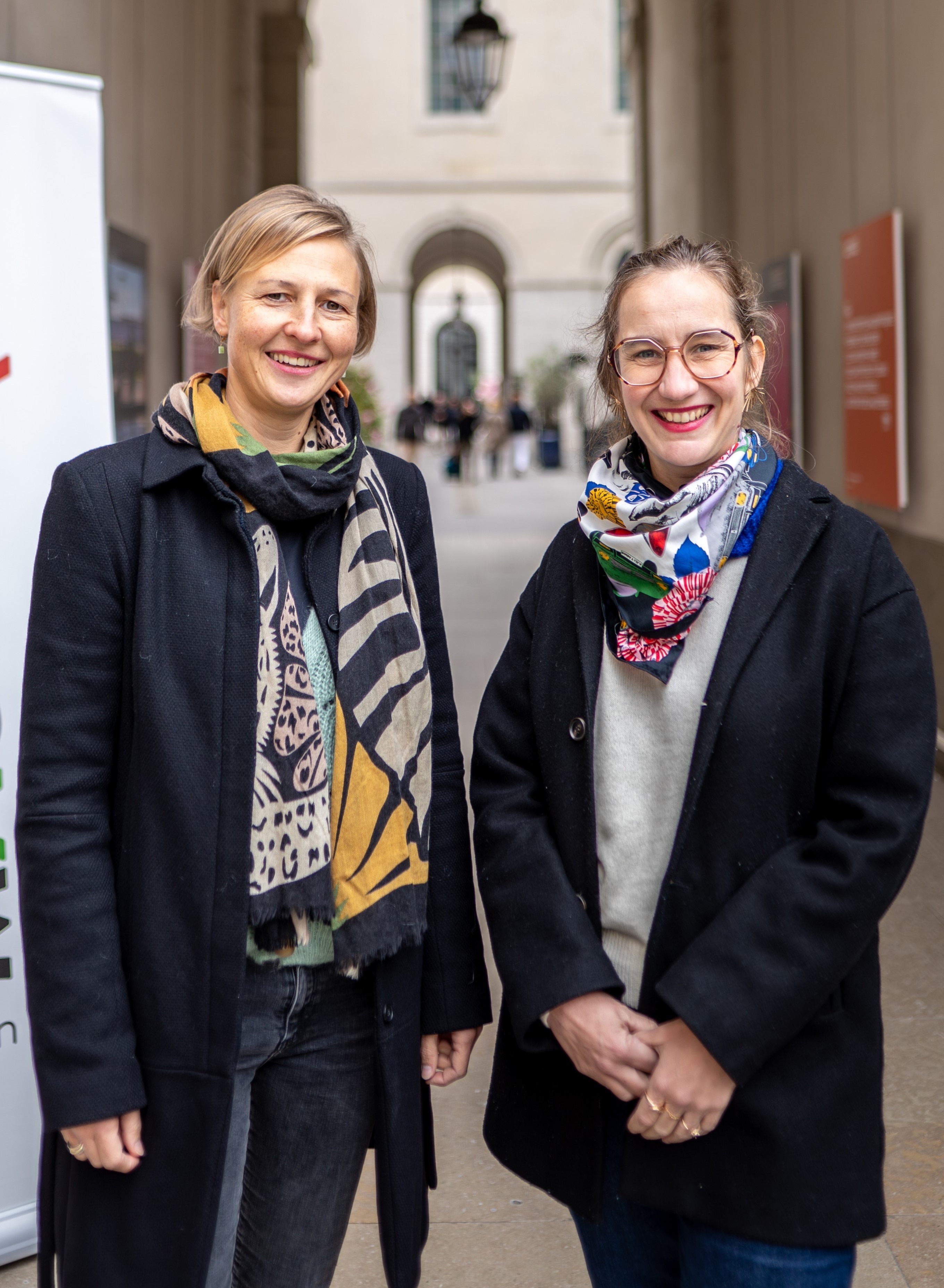 Deux femmes posent côte à côte dans une galerie couverte aux tons clairs. Elles portent toutes deux des manteaux sombres sur des pulls clairs, agrémentés de foulards colorés à motifs autour du cou. La femme de gauche a les cheveux blonds courts et sourit, tandis que la femme de droite porte des lunettes à monture rosée et sourit également en gardant les mains jointes. En arrière-plan, la perspective de la galerie montre des arches et des silhouettes floues, indiquant un espace public ou institutionnel.