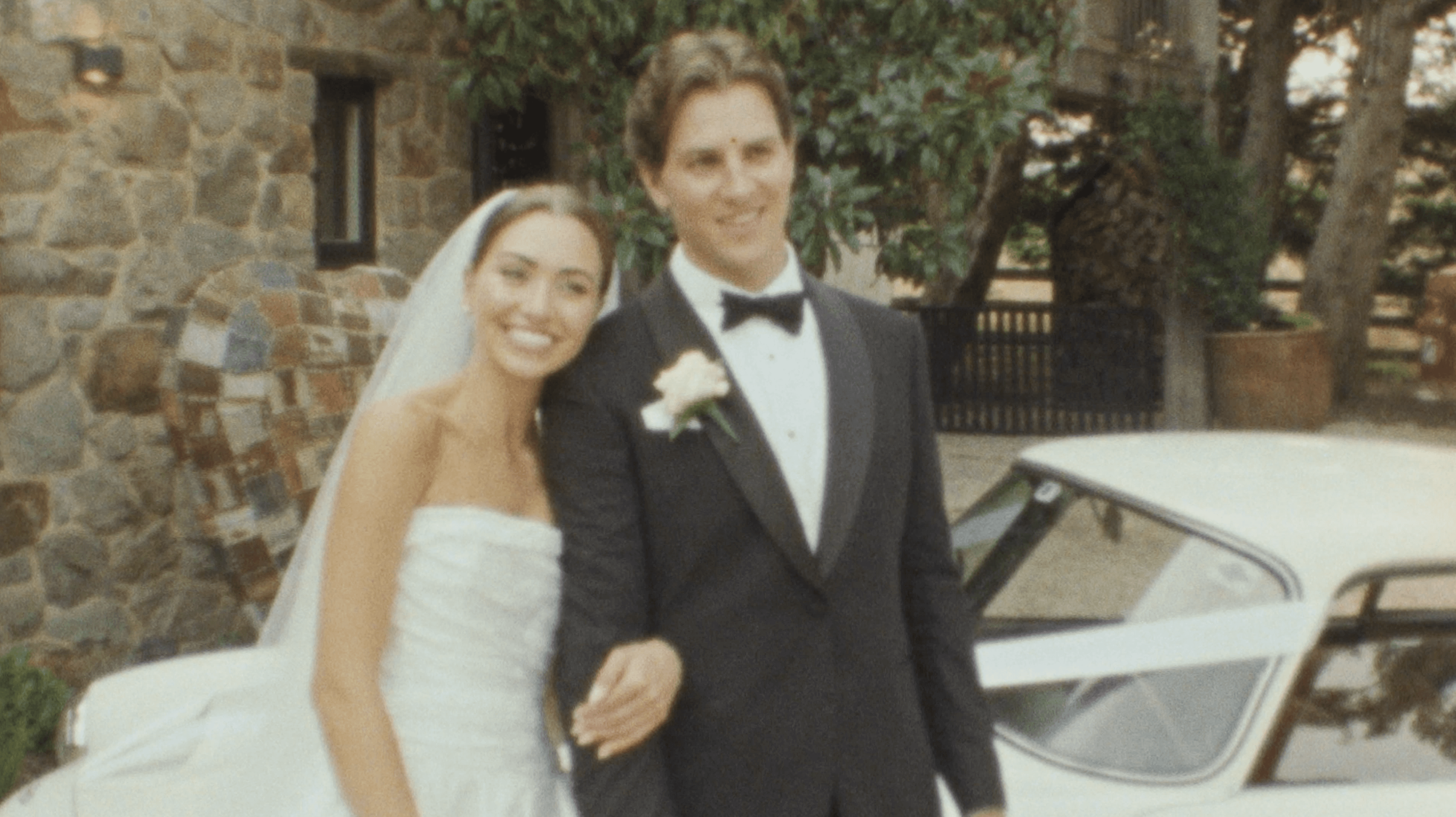 A bride and groom share a kiss under a floral arch with white roses and greenery, as bridesmaids and an officiant in elegant attire stand nearby in a picturesque outdoor setting by a pool.