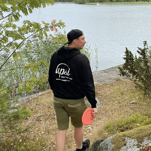 Sami Hammarberg carrying a disc near a stream wearing Upsi hoodie and a Upsi Disc Golf cap