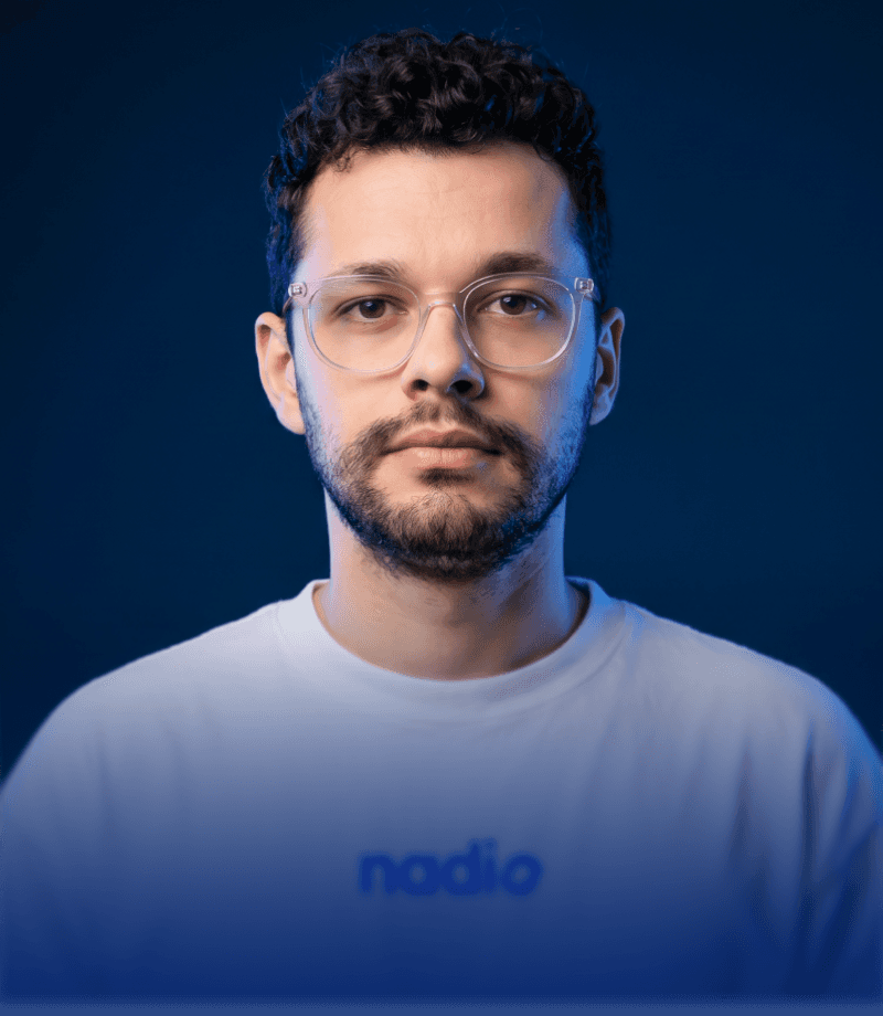 A young man with round glasses, a nose ring, and facial hair looks toward the camera against a dark, softly colored background.