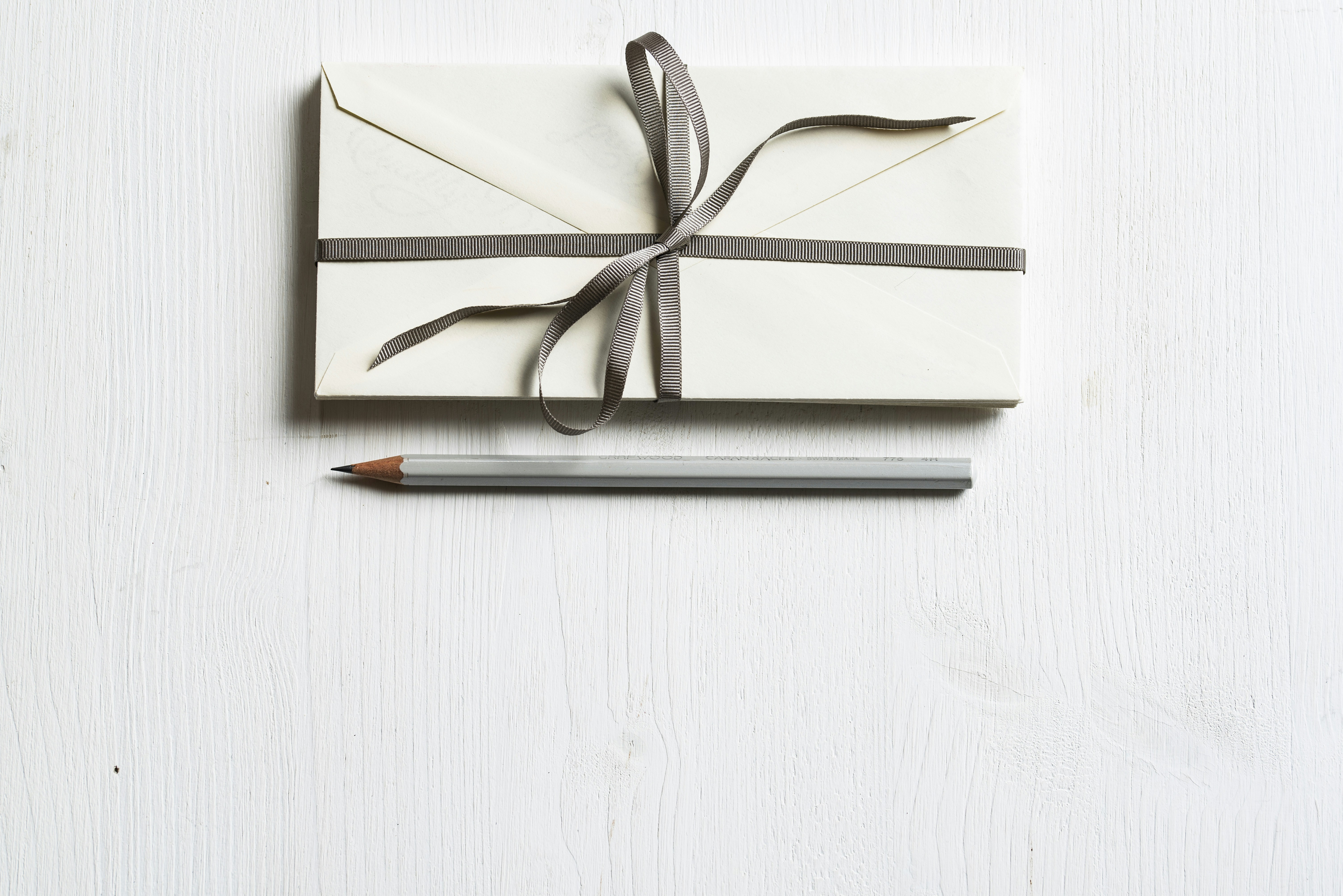 Envelope with dark gray ribbon and a silver pencil on a white wooden surface