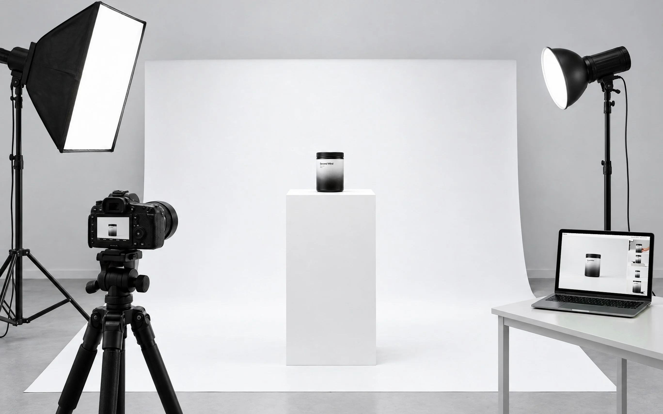 A product shoot: a supplement tub on a white plinth, with a camera on a tripod, studio lights and a laptop showing the shot.