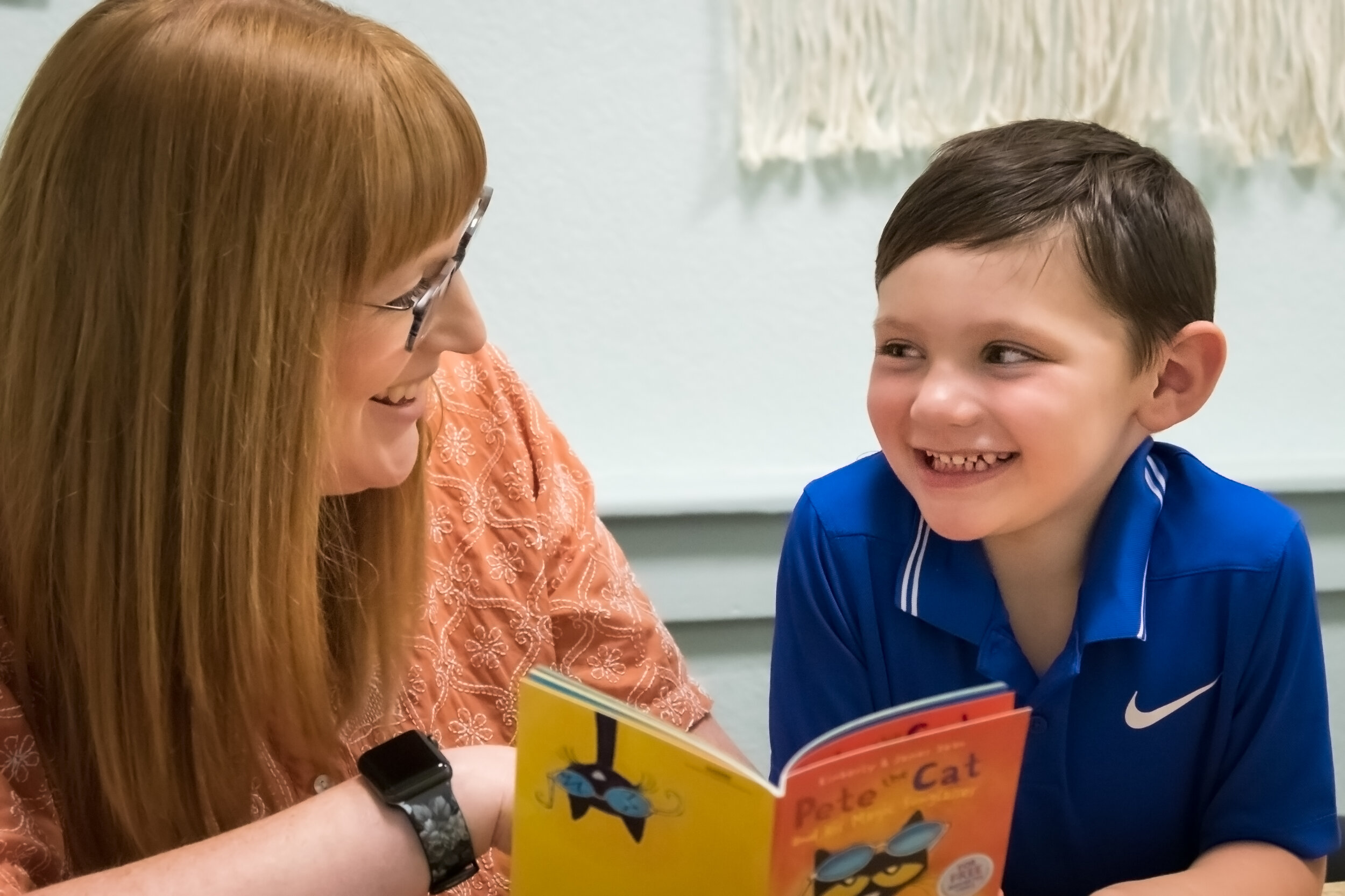 A smiling teacher and young boy share a reading moment, engaging with colorful books in a classroom setting.