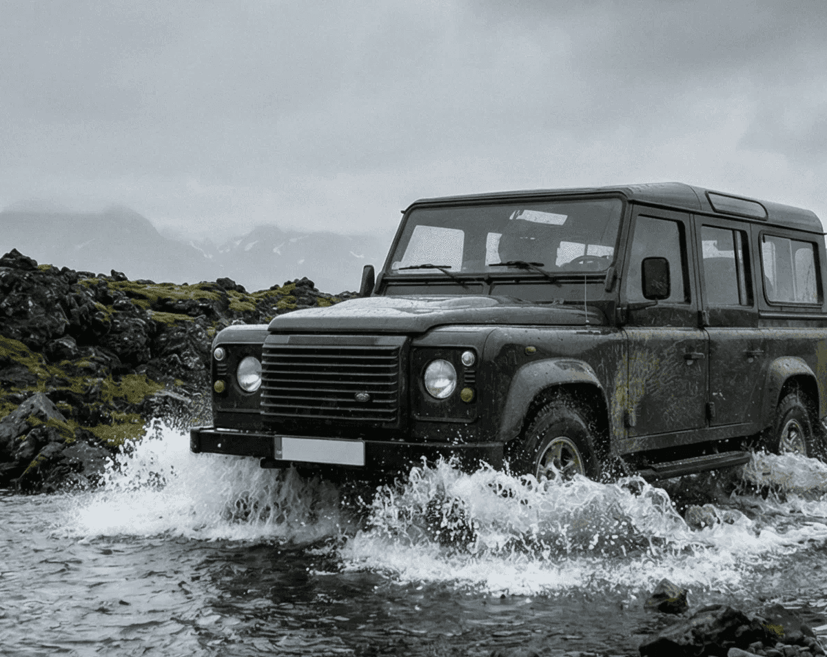 A black Land Rover driving through a river with splashing water, set against a cloudy sky and mossy landscape.