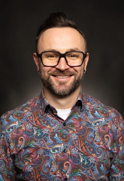 Csaba Horvath, UX Specialist. Smiling man with glasses wearing a colorful paisley shirt, photographed against a dark background, representing Byte/Me Design’s friendly, human-centered approach.