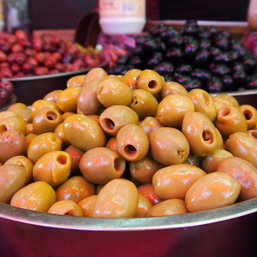 A large bowl of green olives on display at a market, with other types of olives visible in the background.