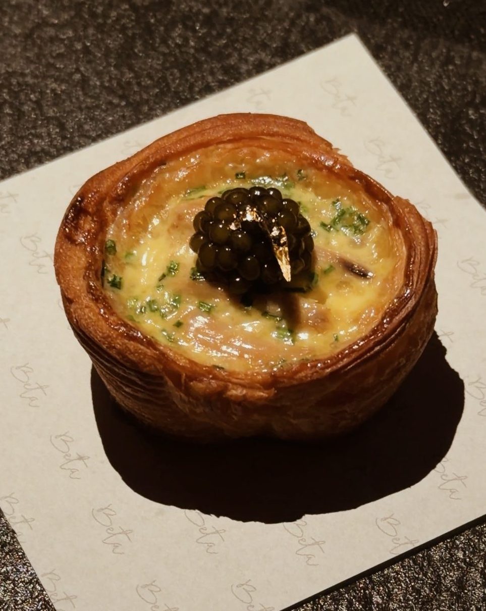 A gourmet pastry topped with caviar and garnished with herbs, served on a branded paper at BETA KL.
