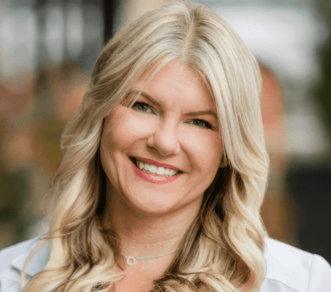 A woman with long blonde hair smiles warmly at the camera, outdoors with a blurred background.