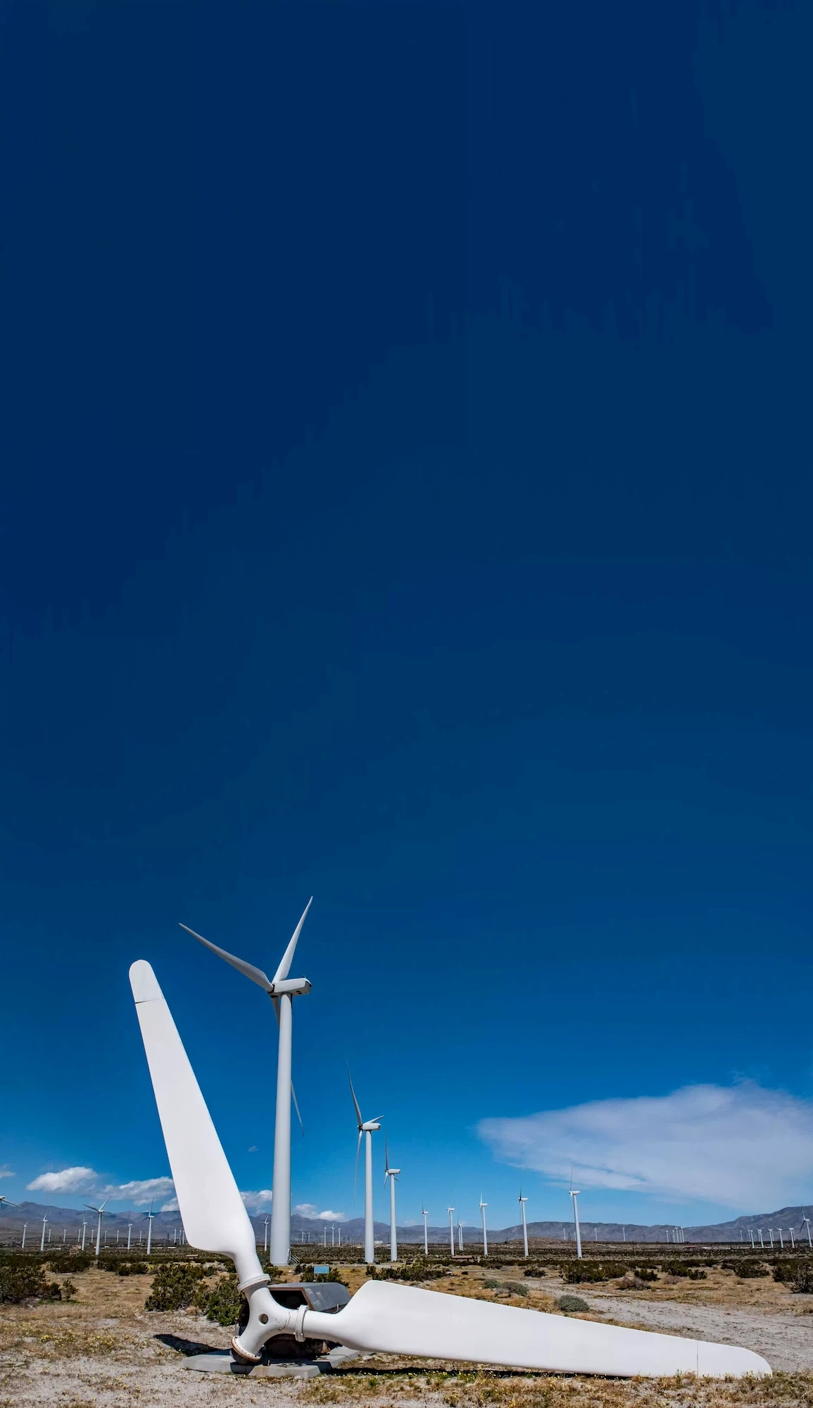 A large broken wind turbine blade lies on the ground in front of several intact wind turbines in a desert wind farm, with clear blue skies and mountains in the background.