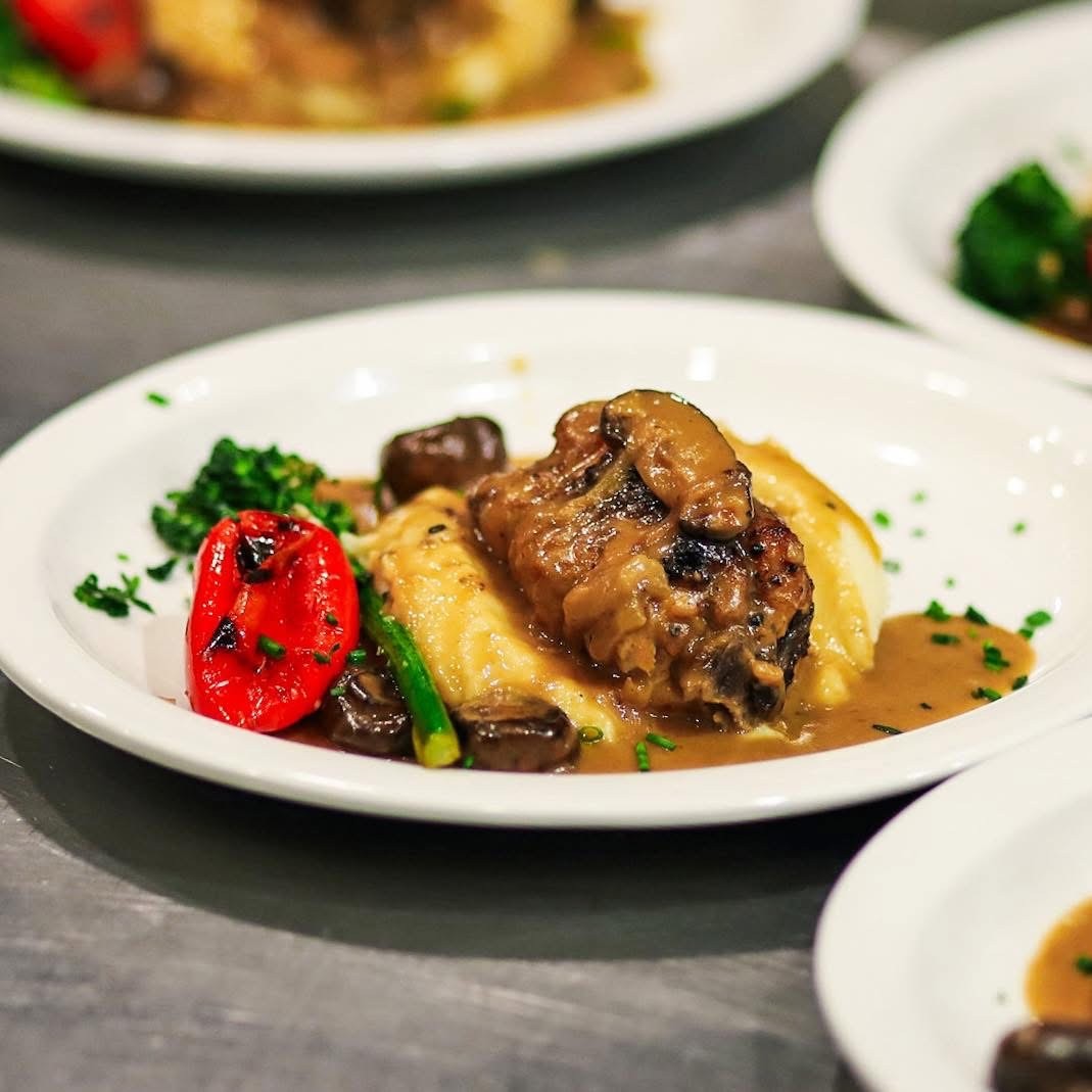 A plated gourmet meal featuring braised short rib served over creamy mashed potatoes, topped with mushroom gravy and accompanied by a roasted red pepper and steamed broccoli.