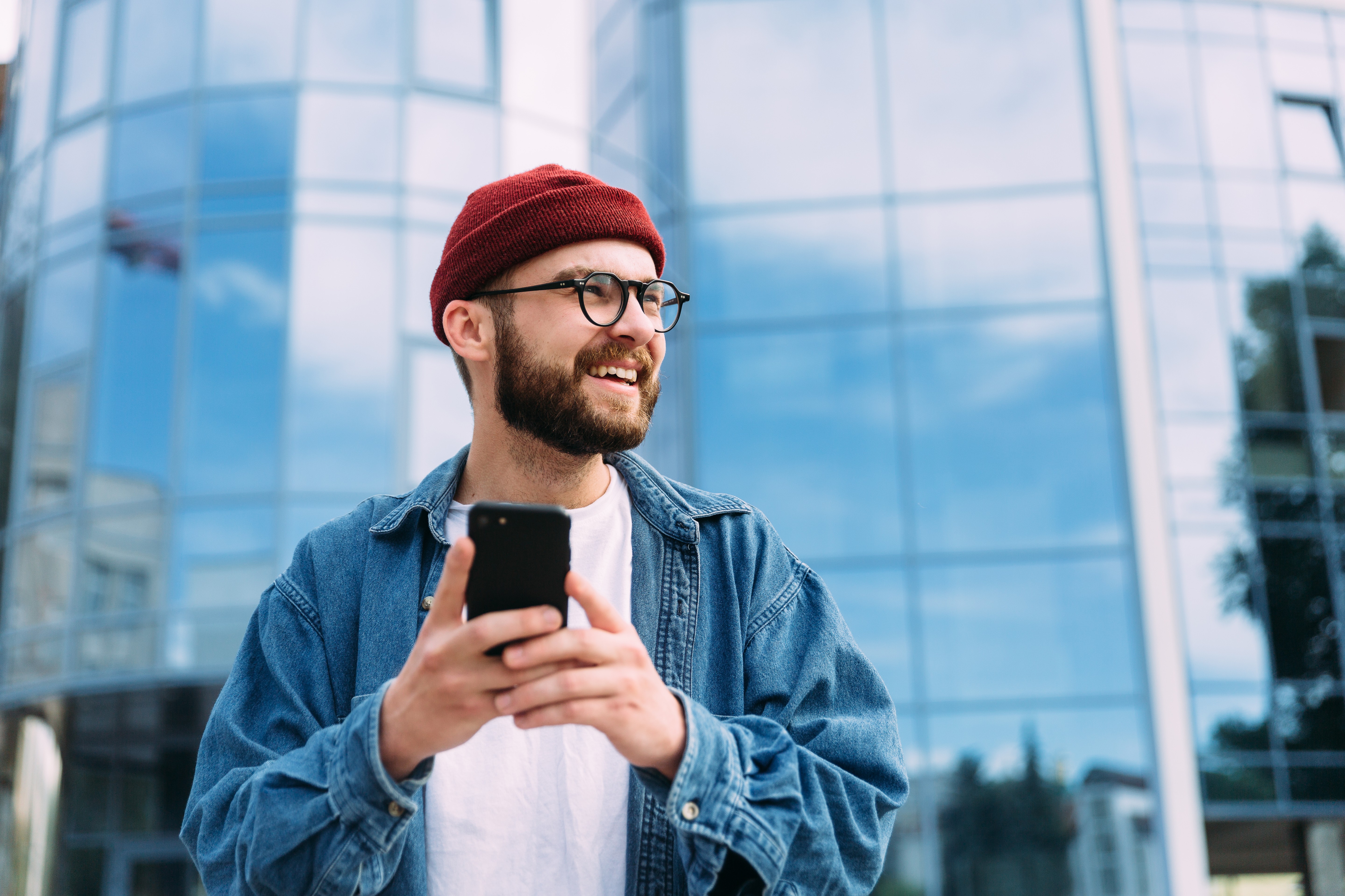 Homem sorrindo com celular na mão, usando gorro vermelho, óculos e jaqueta jeans em frente a um prédio moderno de vidro.