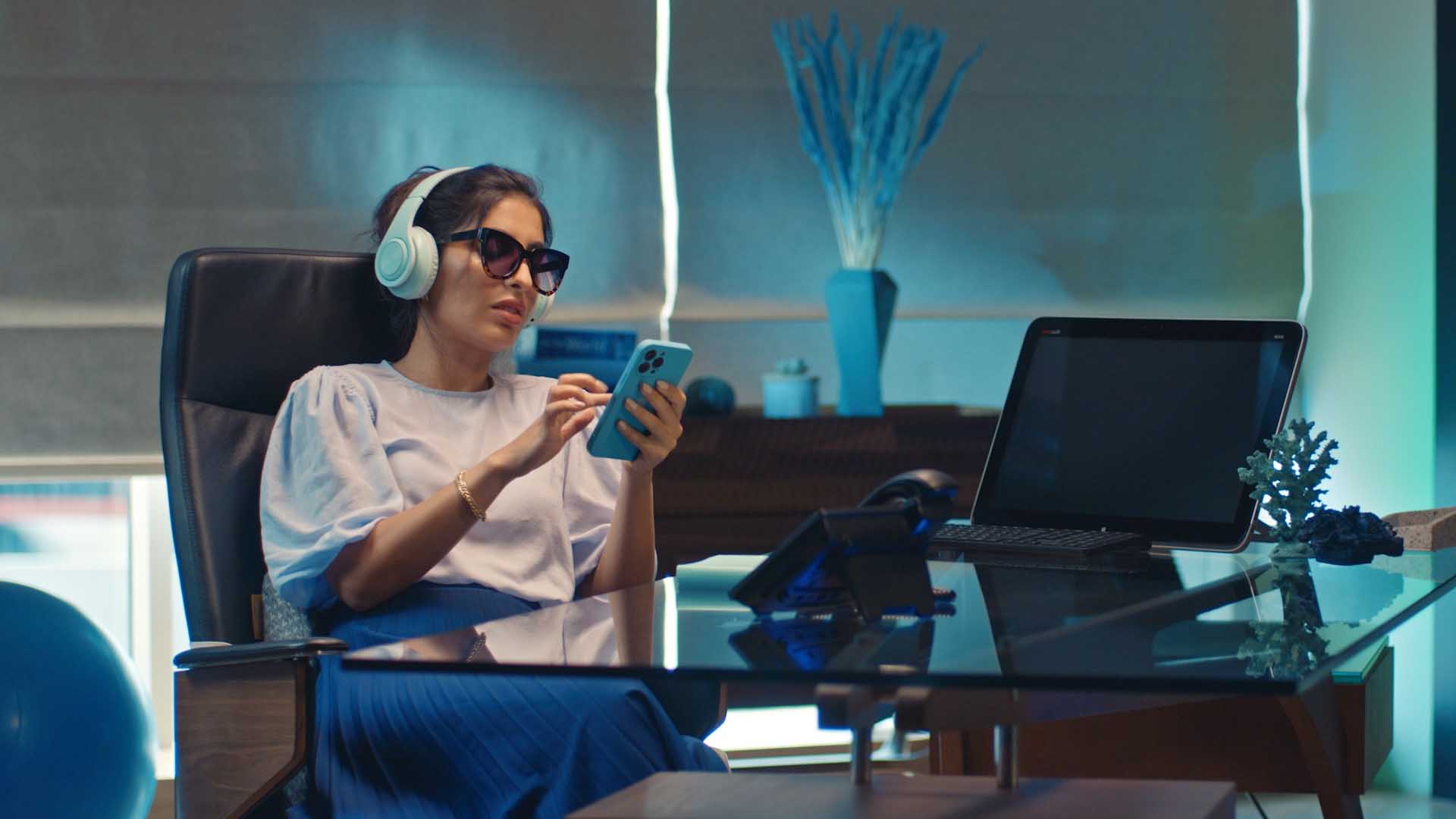 The young woman relaxed in her office chair, with sun glasses and headphones, looking at her phone 