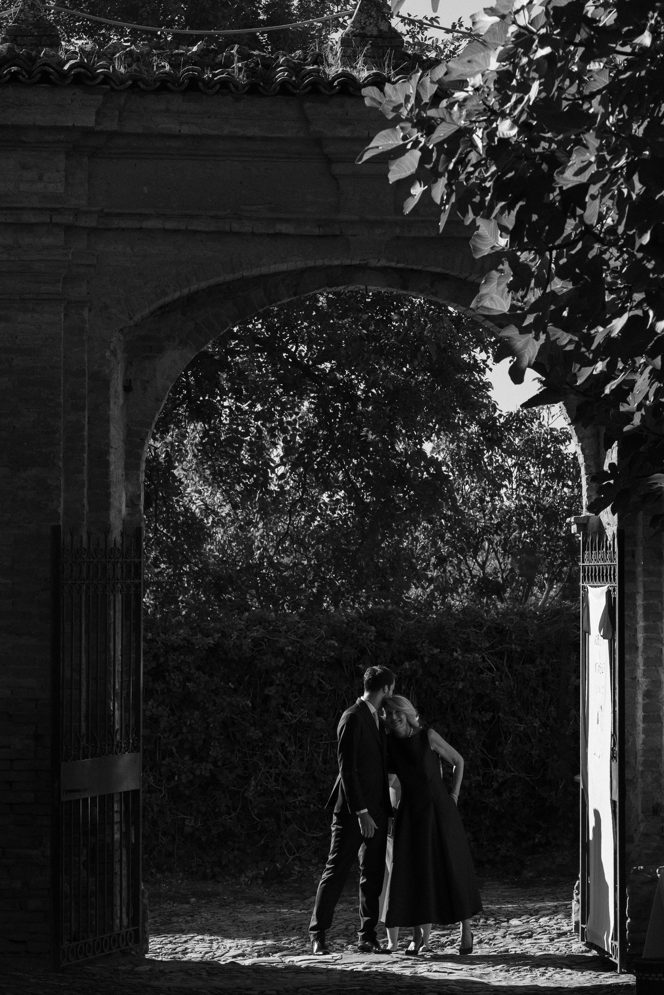 Sposi sotto l'arco in pietra del Castello di Scipione, luce naturale tra le foglie, reportage matrimoniale in bianco e nero