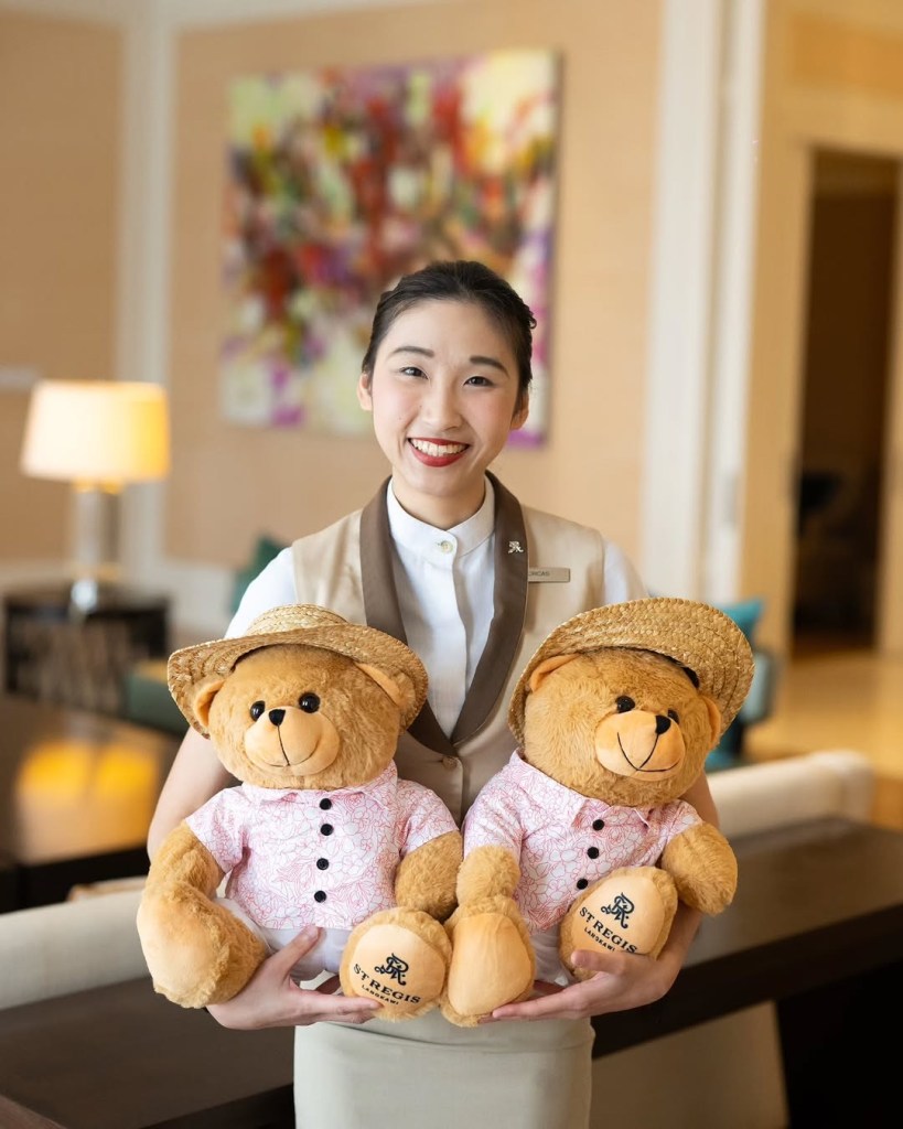 A staff member at The St. Regis Langkawi holding two teddy bears dressed in floral shirts and straw hats, with a colorful artwork in the background.