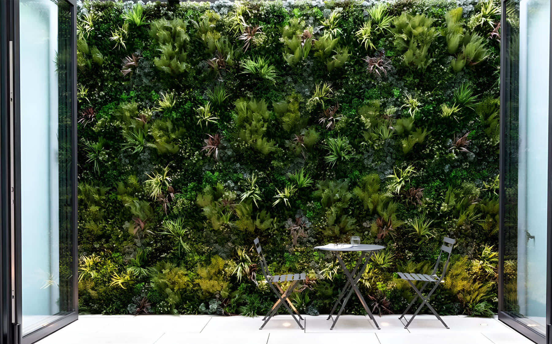 Two large glass French doors open to a patio with a small table and two chairs. In the background, a lush green wall made of VistaFolia artificial plants adds a touch of nature to the outdoor environment.