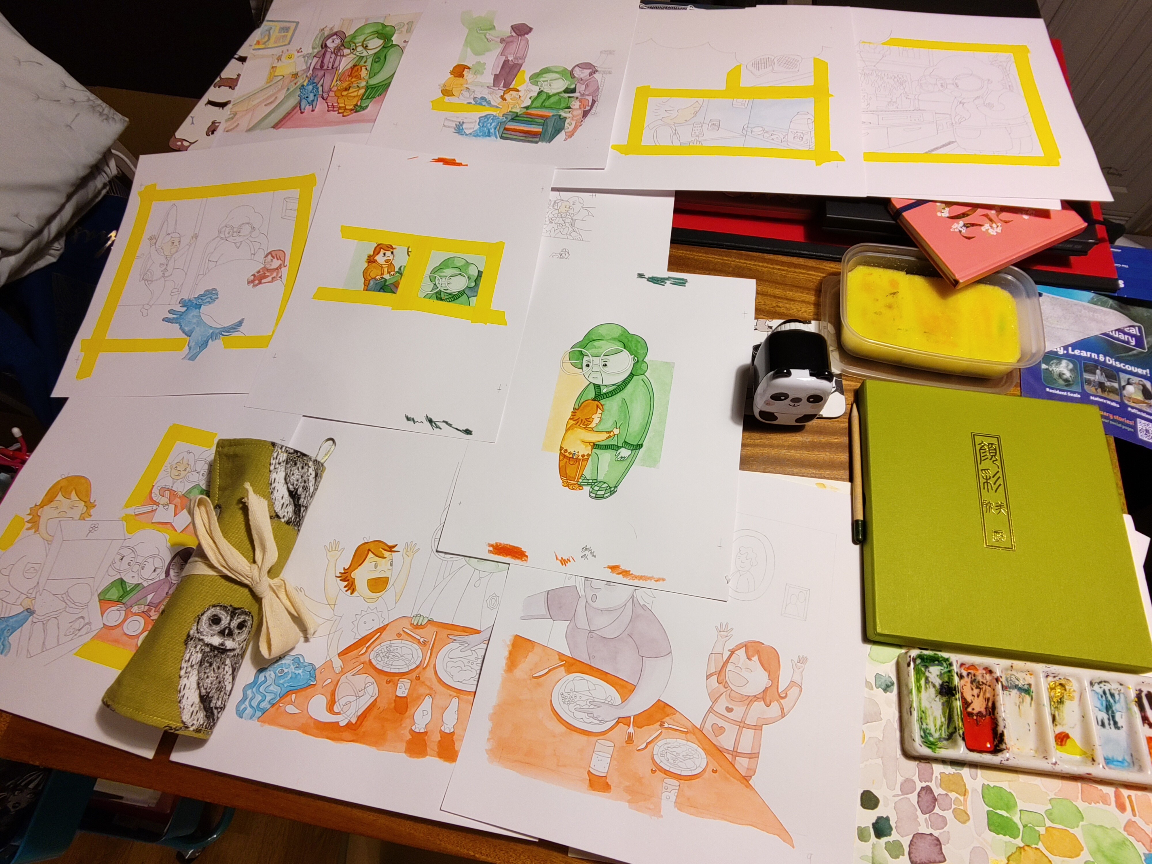 There are many A3 pieces of paper all over a dining room table. They show many illustrations of the book all at different stages, some are pencil sketches, other are part painted. Some have washi tape stuck down which is used to mask off the area for painting. There is a role for pencils and a box of paints. 